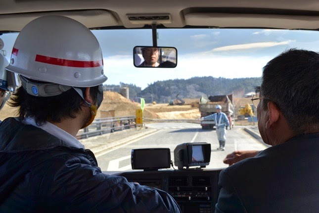 ヘルメットをかぶった二人の大人が車の中から前方の工事現場を見ており、道路には反射ベストを着た作業員が立っている、車内から写した写真