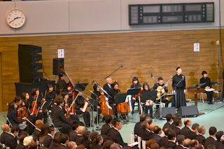 黒の服を着用した楽器を構えた楽団と追悼式に訪れた人々が並べられた椅子に座っている写真