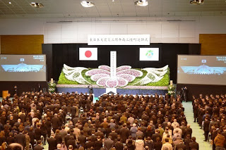 祭壇の両脇に大きなスクリーンが用意され多くの人々が整然と並び立ち黙とうを捧げているところを後ろから写した写真