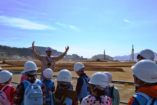ヘルメットを被った子供たちが柵の向こうの工事現場を見ながら説明を聞いており、男性が両手を広げて現場説明をしている写真
