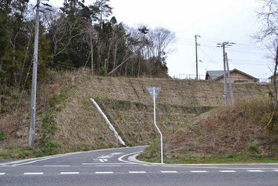 坂道の入口にある交差点で、奥の草地の法面上に家屋の屋根が見えている写真