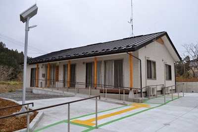 建物の前に点字ブロックと、手すり付きのスロープが設置され、軒下にオレンジ色の柱が並ぶ、白壁の平屋建ての集会所の建物外観の写真