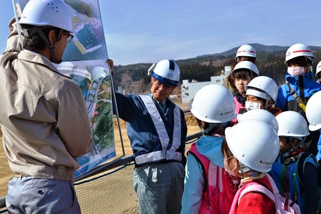 ヘルメットを被った男性が大きなパネルを持ち、もう一人の作業服を着た男性が、子供たちに建設計画について説明している写真