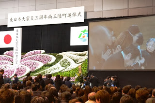 多くの人々が参列する東日本大震災三周年南三陸町追悼式の会場で、祭壇の前に整然と並び手を合わせている様子が大きなスクリーンにも映し出されている写真