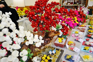 白や赤、ピンクのシルクフラワーの花鉢が並んでいる前にコサージュが並べられている写真