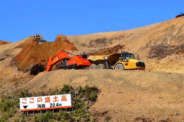 「ここの盛土高 海抜20.4メートル」と書かれた白地に赤文字の看板の前に、赤いショベルカーと黄色いダンプトラックが作業中の、盛土工事の様子を示した写真