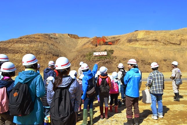 ヘルメットをかぶった子供たちと大人たちが盛土の方角を見上げており、その上方には重機があり、看板を指差す子供の姿も見られる、土木工事の見学中の写真