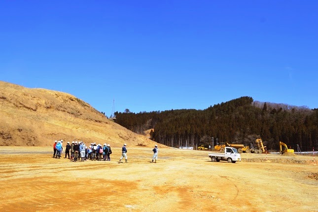山を崩してできた広い平地に子供たちを含む見学者の集団が集まっており、周囲には黄色い建設車両と山林が見える、造成地での見学の写真
