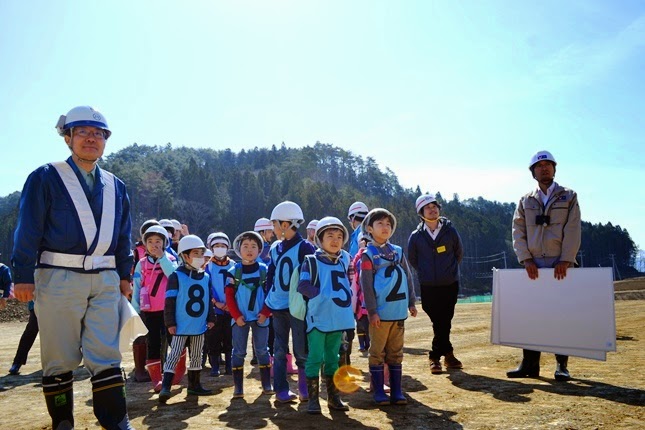 青いビブスを着てヘルメットをかぶった子供たちが、建設現場で説明を受けるために整列して真剣な表情で前を見つめている様子の写真