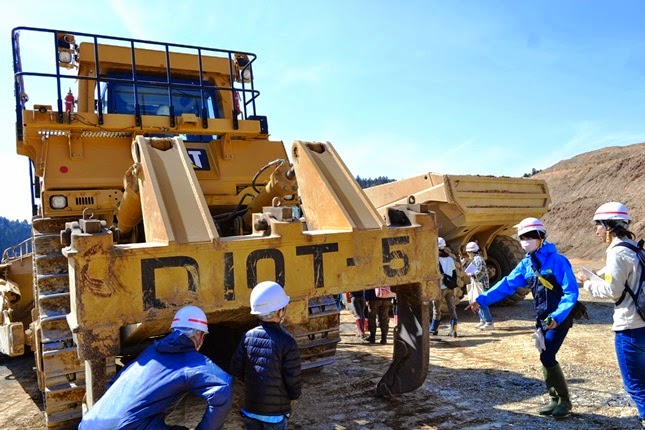 子供たち数名がヘルメットをかぶり、巨大な建設機械の後方に集まり、ブルドーザー の後方に装着された「リッパー」という機械を見学している写真