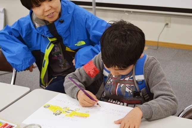 屋内で、男の子が工事車両の絵を描き黄色く塗っている絵などが描かれた写真