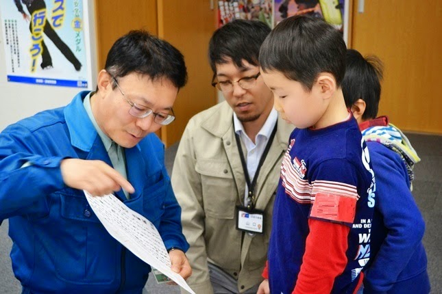 室内で青い作業服の男性が紙を手に持ち、指で指し男の子に話している写真