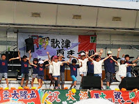 「歌津夏祭り」と書かれた横断幕が掲げられたステージの上でたくさんの子供たちが並んで両手を上に広げたポーズをしている写真