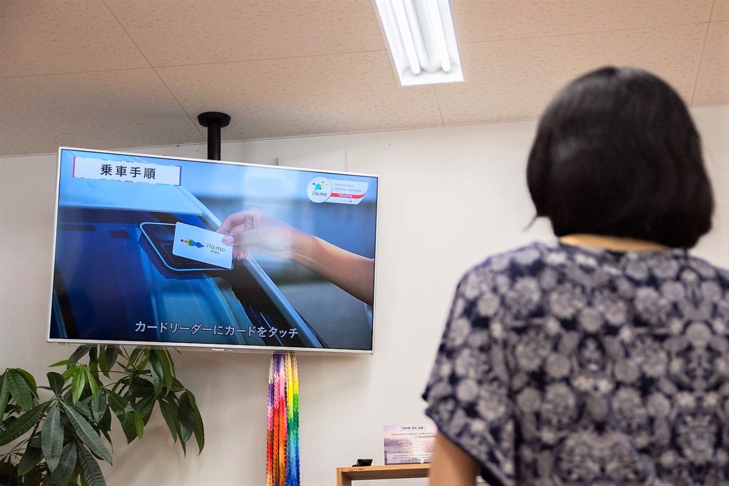 室内で、乗車手順を説明する映像が映るテレビ画面を、背を向けた女性が見ている写真