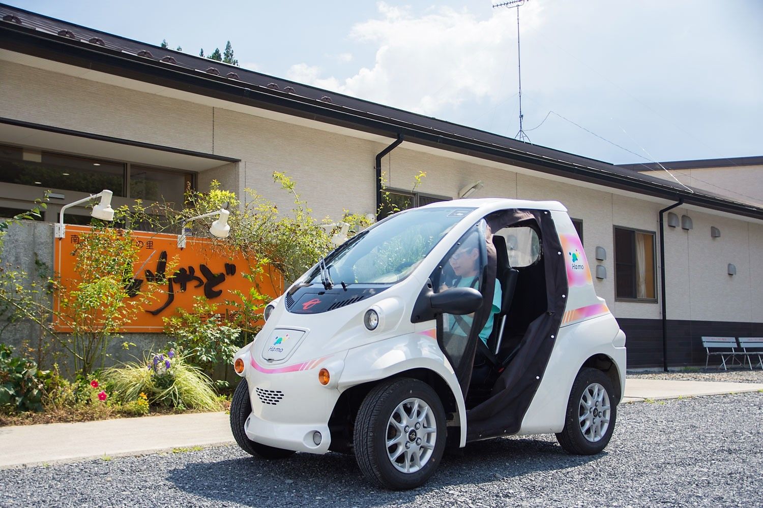 「いりやど」と書かれたオレンジ色の看板がある建物の前に、白い電気自動車を停車させている女性の写真