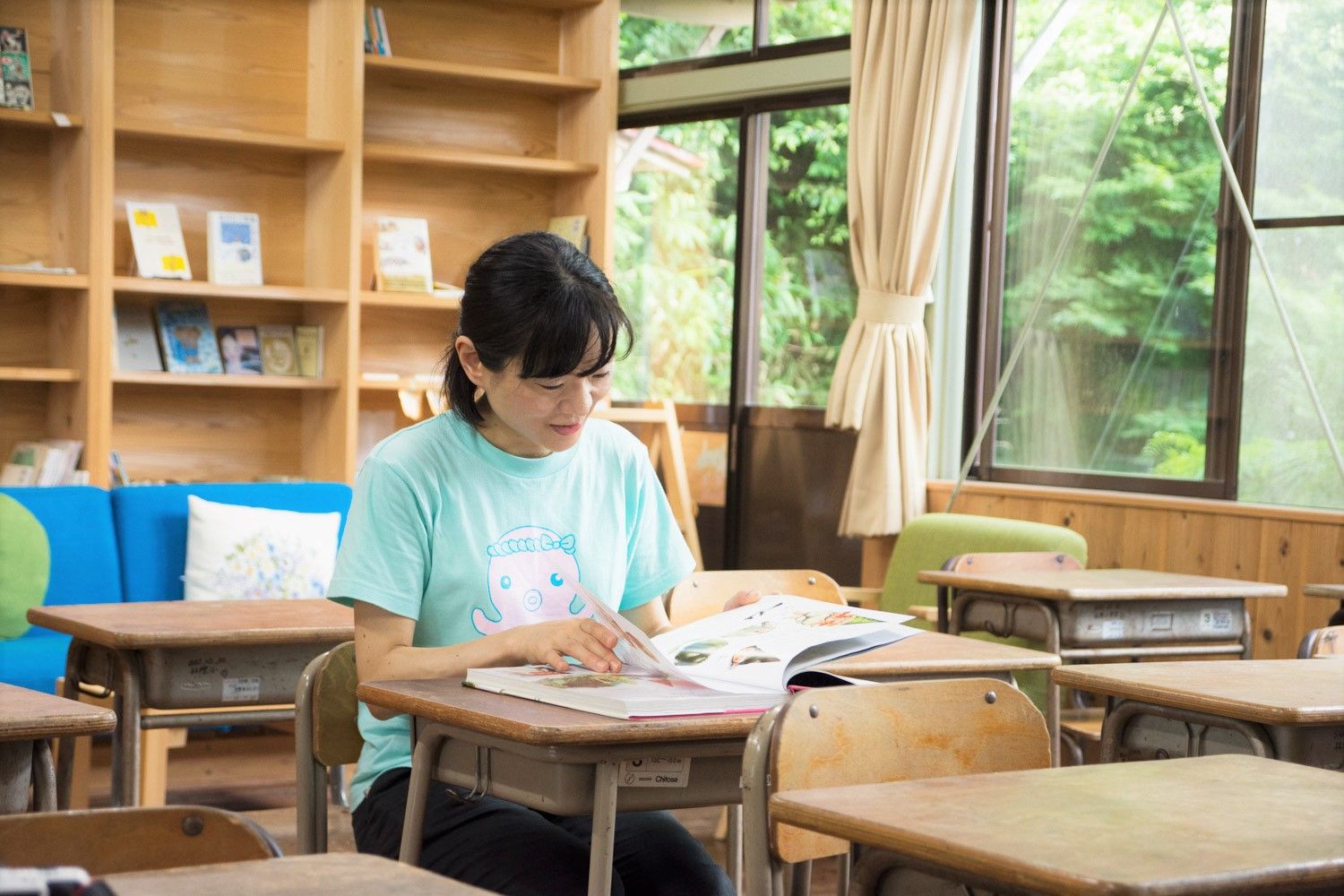 木の本棚と窓際に囲まれた明るい教室で、机に座って本を読んでいる女性の写真