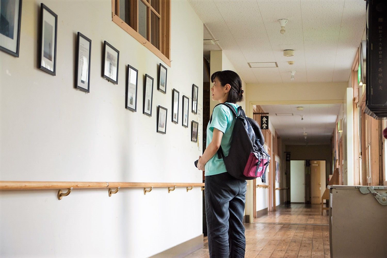 リュックを背負った女性が、廊下の壁に並んだ額縁の写真を立ち止まって見ている写真