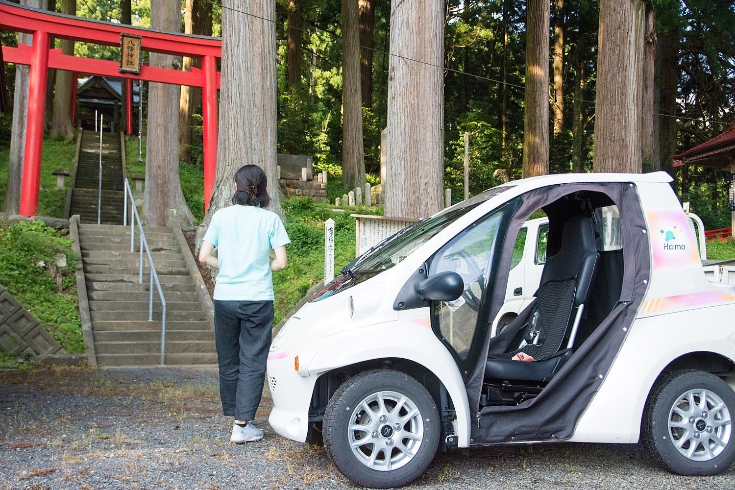 森の中の神社に続く石段の前で赤い鳥居を見上げている女性と、その隣に止まっている小型電気自動車の写真