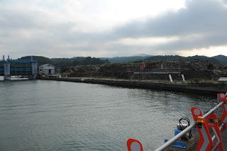 海に面した岸壁の向こうに、積まれたがれきの山が広がり、左手には青い鉄骨の建物と倉庫、手前には工事用の柵が並んでいる様子の写真