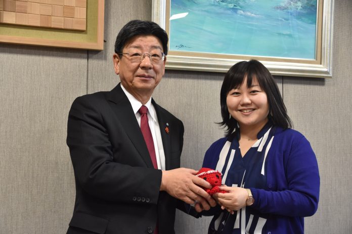 赤いねじり鉢巻きを巻いたタコのキャラクター『オクトパス君』の小さなぬいぐるみを、佐藤町長と女性リポーターが一緒に持ち、笑顔で写っている写真