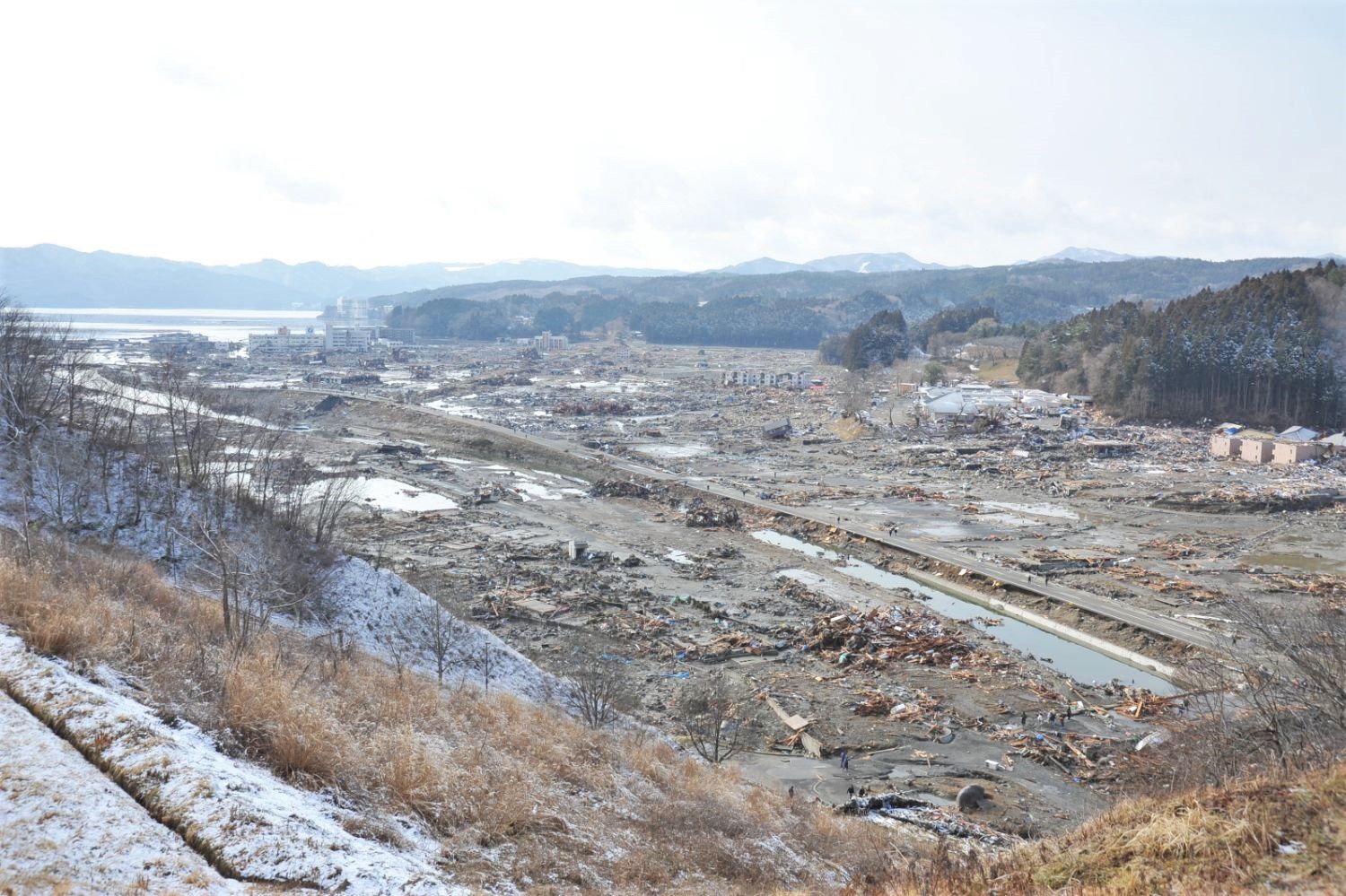 雪がうっすらと積もる寒空の下、津波によって建物や道路が押し流され、木材やがれきが広範囲に散乱し、かつて街であった場所がほぼ更地と化して川の流れだけが残る、東日本大震災直後の被災地の写真