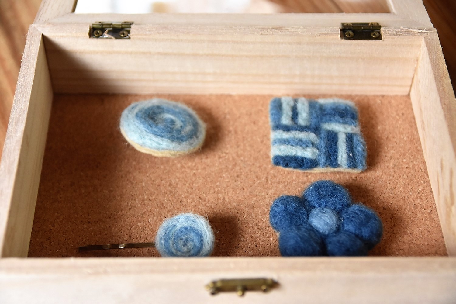 コルクシートがひかれた木の箱の中に羊毛を藍で染めて作った渦巻やお花や四角の形をしたブローチやヘアピンが4つ並べられている写真