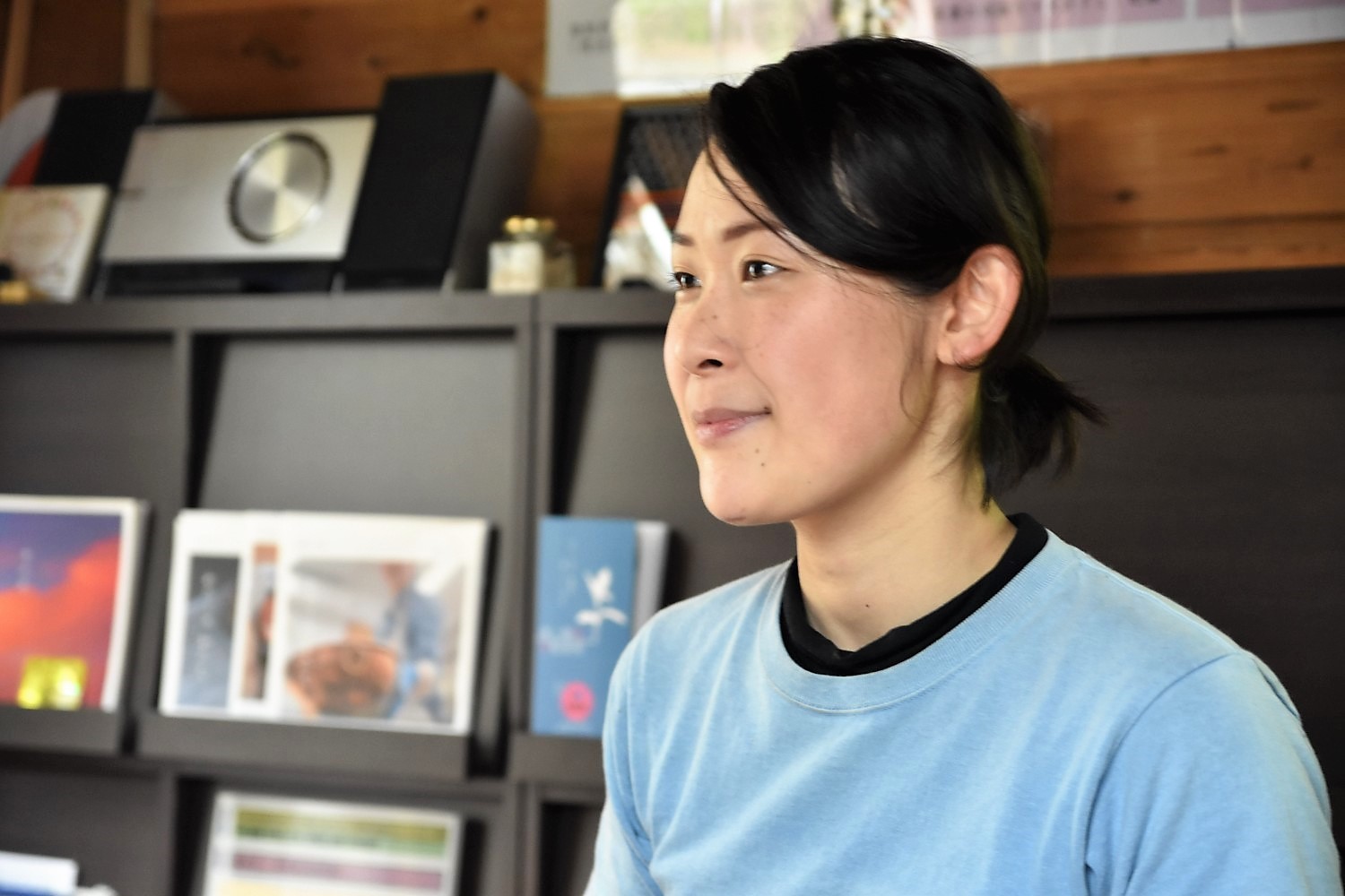 ディスプレイラックに本が飾られている部屋で座っている水色のTシャツを着た中村さんのアップの写真