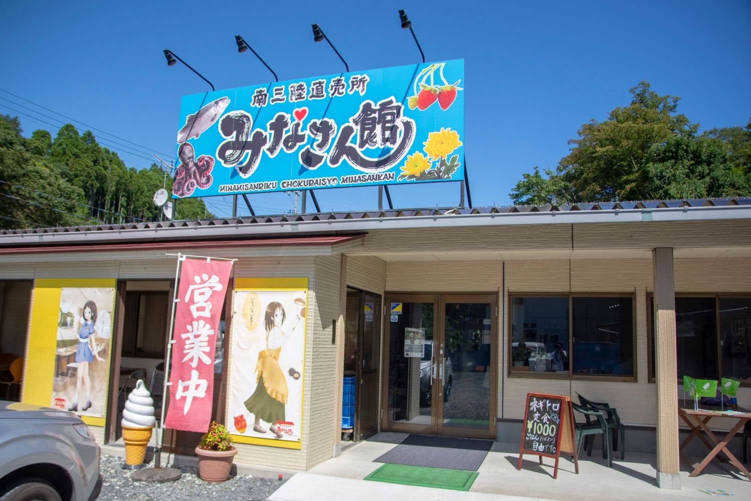 「南三陸直売所みなさん館」の文字とイチゴやタコ、魚や黄色の花などのイラストが描かれた水色の看板が設置され、営業中と書かれたのぼり旗が設置された建物外観の写真
