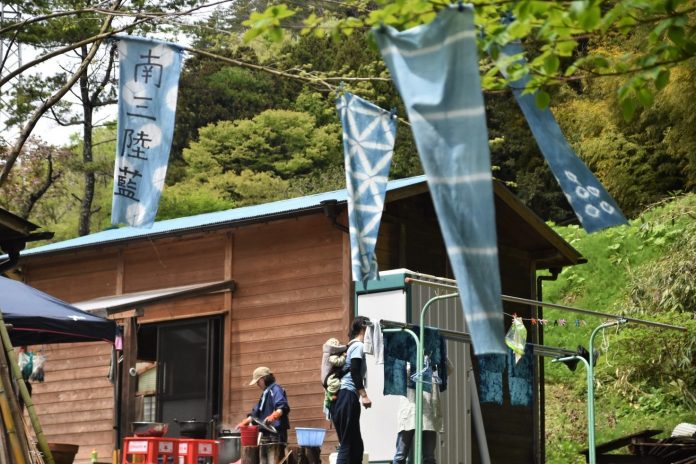 木々の緑に囲まれた山あいの場所に建つ茶色い木造小屋の前で、青い藍染めの旗が風に揺れ、子どもを背負った女性や作業する方々がいる藍監査室の写真
