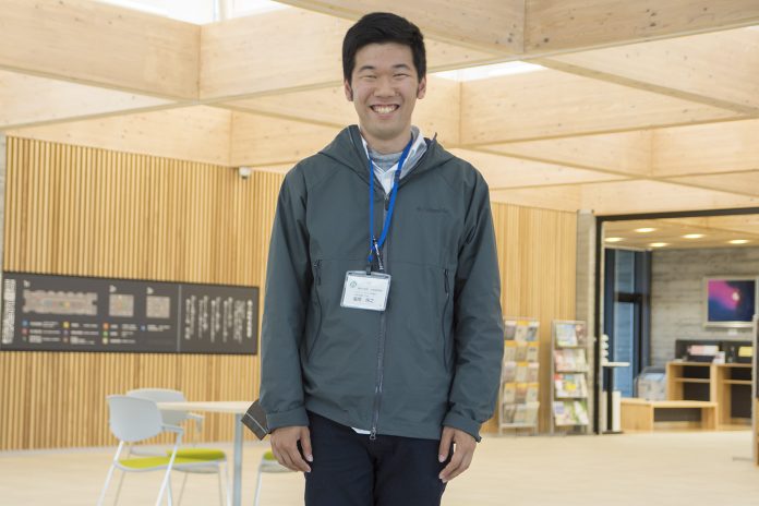 木の梁が見える開放的な屋内で、グレー色のジャケットを着て青いネームストラップを下げた男性が正面を向いて笑顔で立っている写真