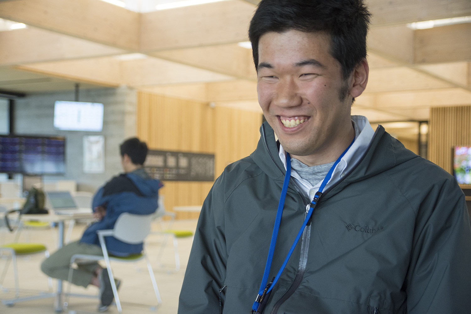 背景には別の人物が椅子に座って作業しているスペースで、グレー色のジャンパーを着た男性を右斜めからアップで撮影した写真