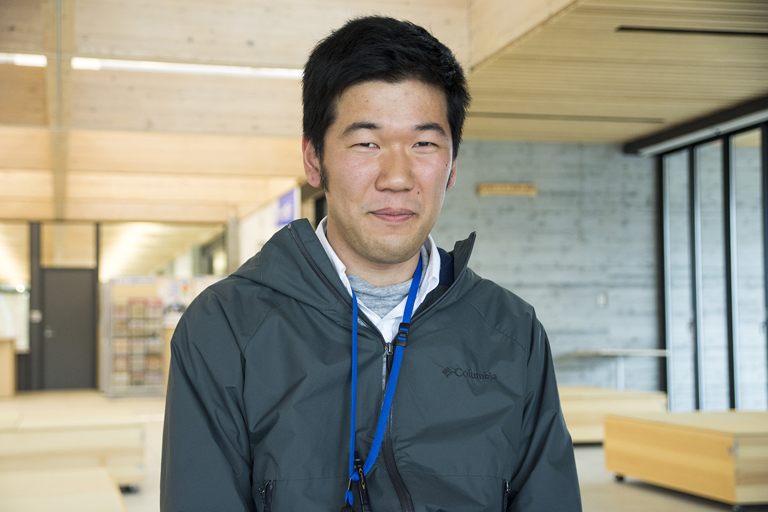 木の梁とガラス窓がある現代的な建物の内部で、男性がやや控えめな表情でカメラを見つめている写真