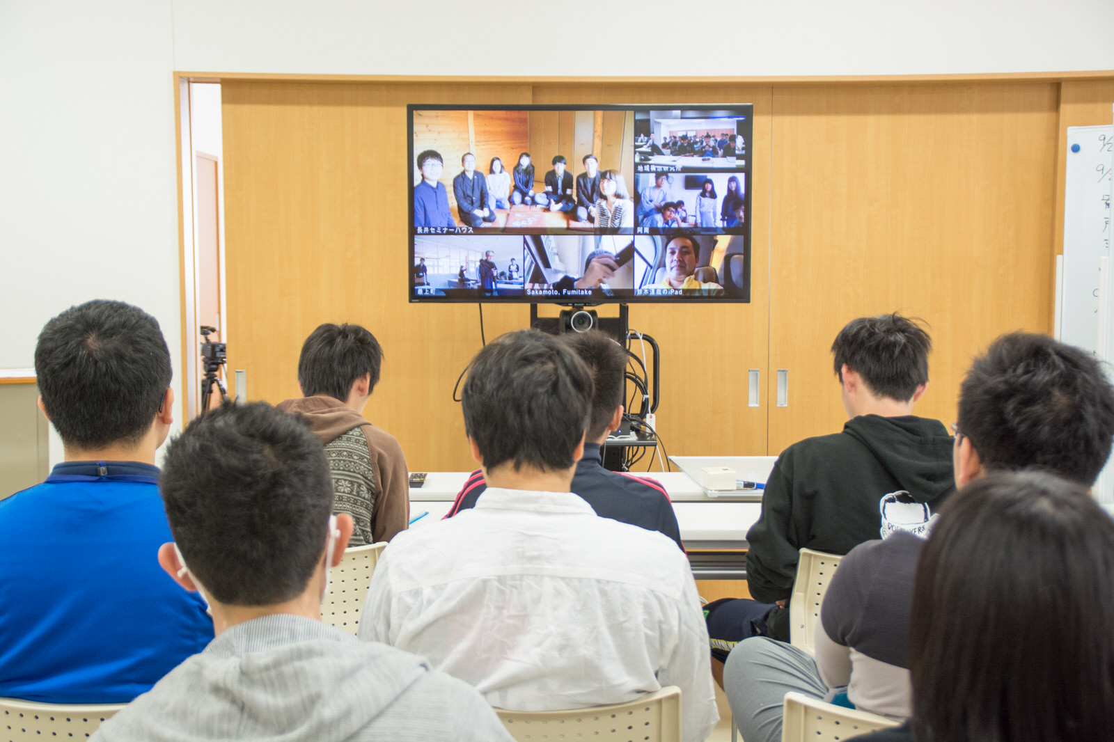 教室で8人の学生たちがモニター画面に映る他地域の人達とのオンライン会議を行っている後ろ姿の写真