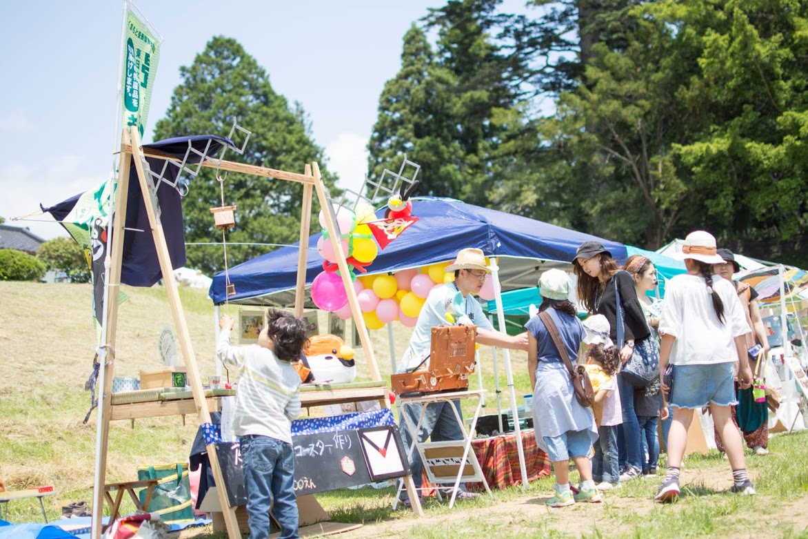 カラフルなテントと風船が並ぶ広場で、子どもや大人たちがテントの前に集まって楽しそうに過ごしている様子を写した写真