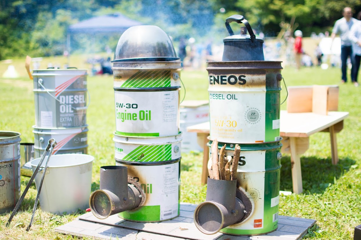 ステンレス製のボウルや薬缶が上に置かれた煙突から煙が出ており、芝生の上に空き缶やオイル缶を再利用して作られた手作りのロケットストーブが2台置かれている写真