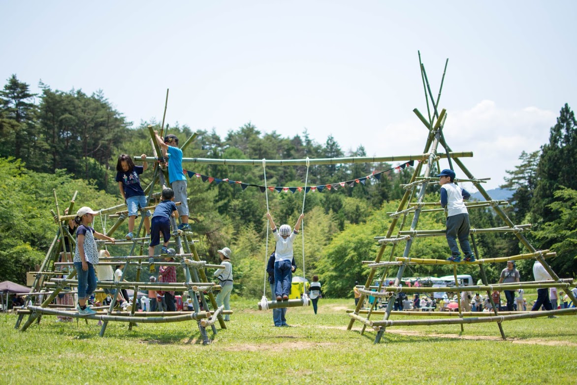 芝生広場に設置された、竹で組まれた三角形の大型ジャングルジムのような遊具で、子どもたちが元気に登ったり遊んだりしている写真