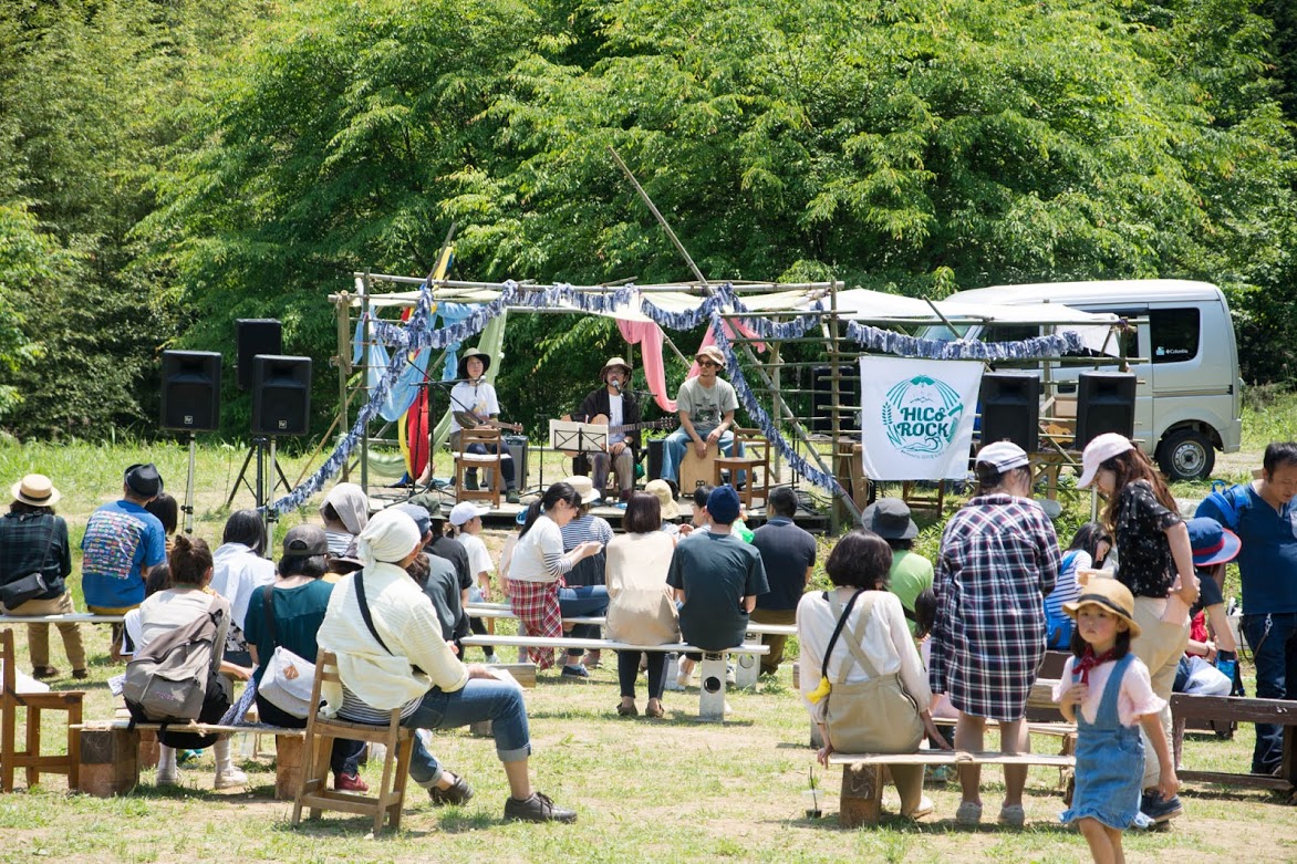 竹で作られたステージの上で複数の演奏者が音楽を披露し、その前に観客が木製ベンチに座って演奏を楽しんでいる写真