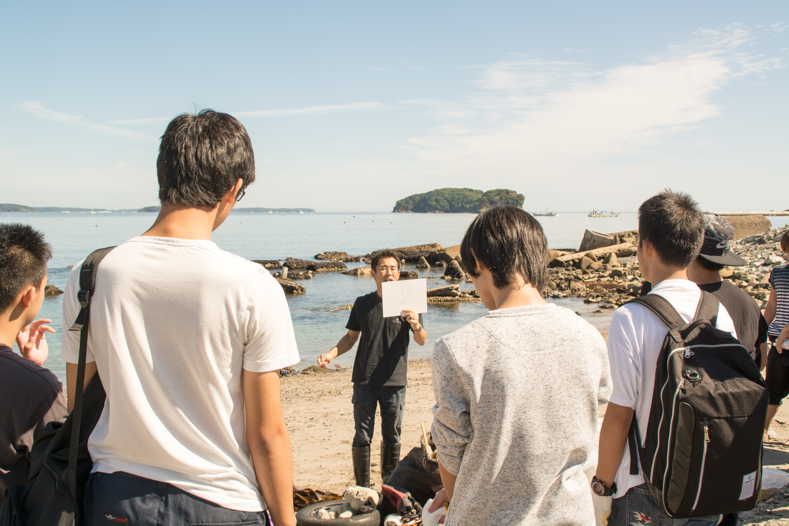 海岸で紙を掲げて説明している一人の男性の話を学生たちが真剣に聞いている後ろ姿の写真