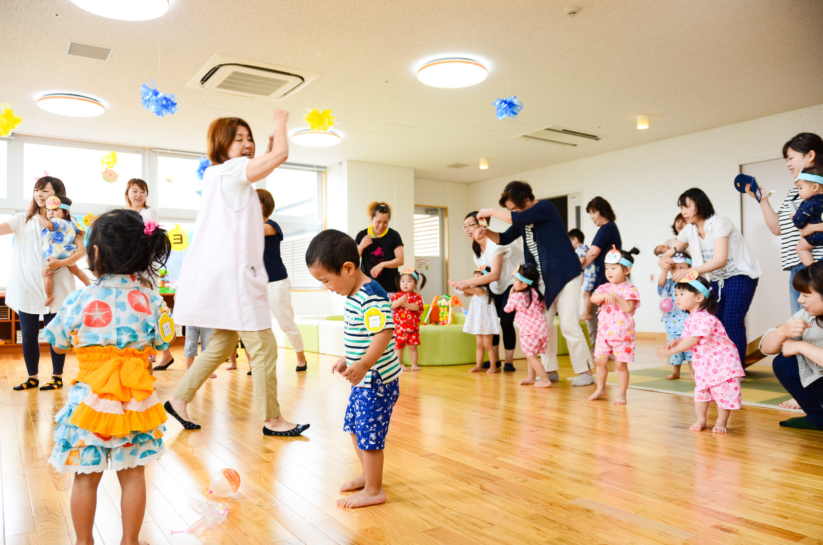 天井から青と黄色の飾りが複数吊り下げられた部屋で、スタッフや浴衣を着た子どもたち、保護者が輪になって盆踊りを楽しんでいる写真