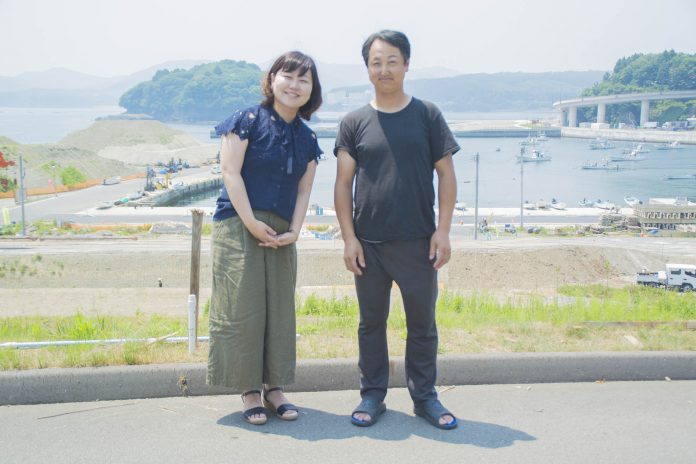 リポーターの女性と佐々木さんが、青い海と船の浮かぶ港、そして湾に架かる橋を背景に舗装された道路上で並んで立っている写真