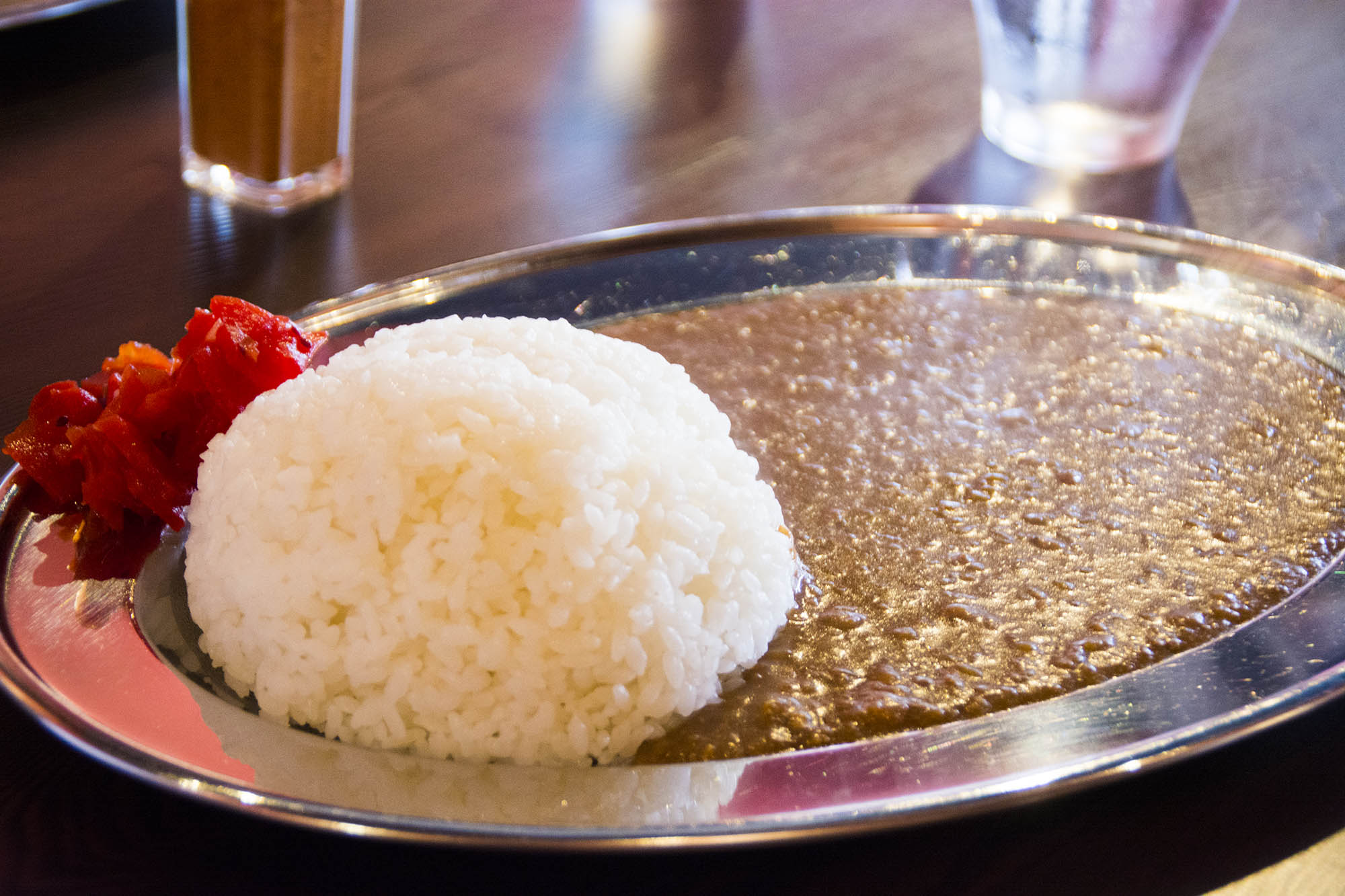 銀色の楕円形の皿に、丸く形作られた白ごはんの横に赤い福神漬けが添えられ、カレーソースが皿の半分以上に広がっているカレーライスの写真