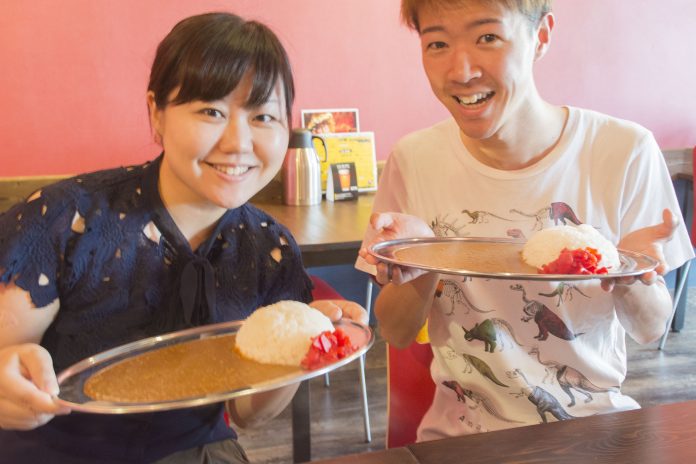 リポーターの女性と佐藤さんがそれぞれ福神漬けが添えられたカレーとライスが盛り付けられた銀の皿を両手で持ち、笑顔でカメラに向かって差し出している写真