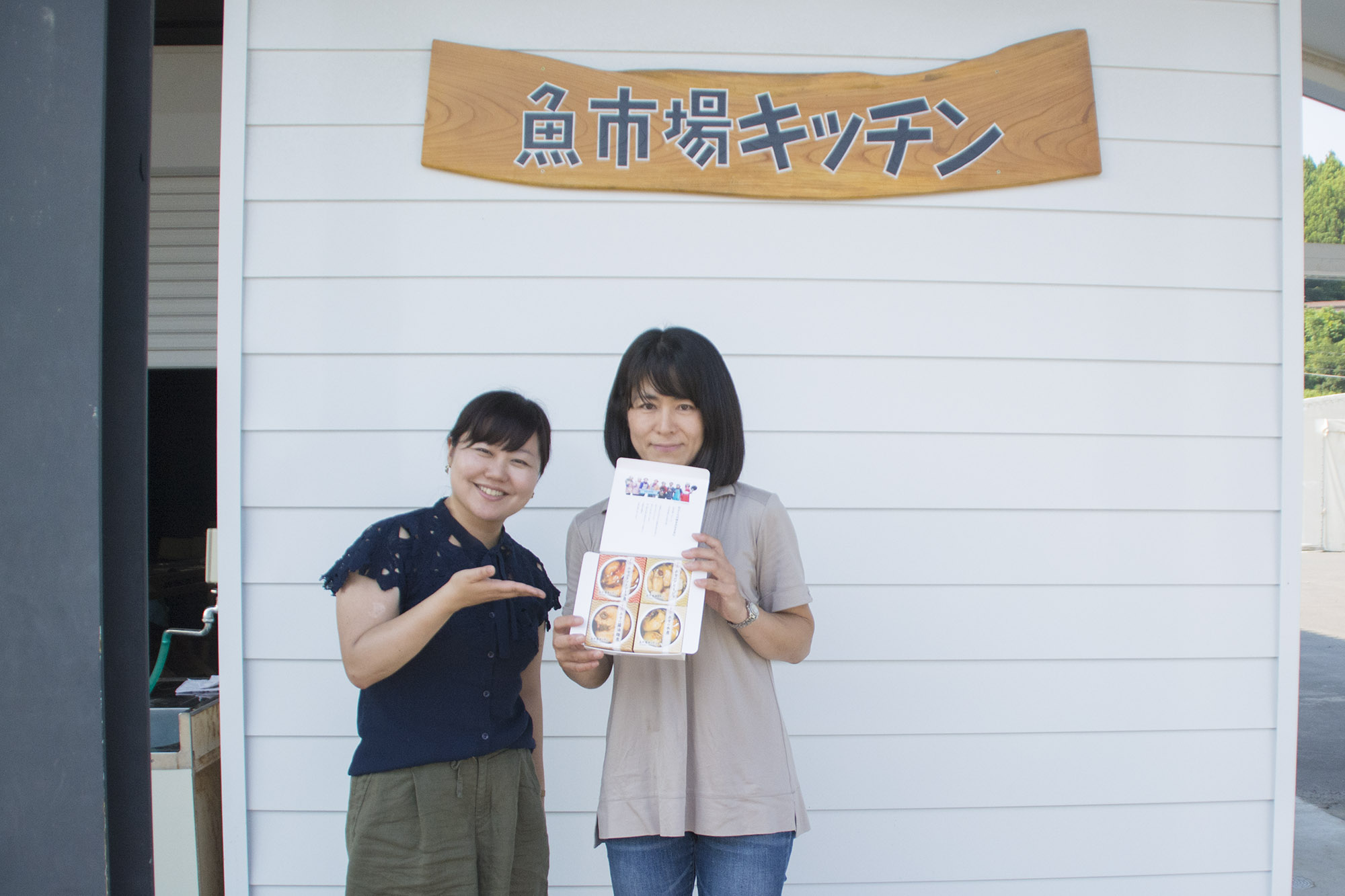 「魚市場キッチン」と書かれた木製の看板が掲げられた白い壁の前で、リポーターの女性が、笑顔で右に立った女性が手に持った商品の詰め合わせセットを紹介している様子の写真