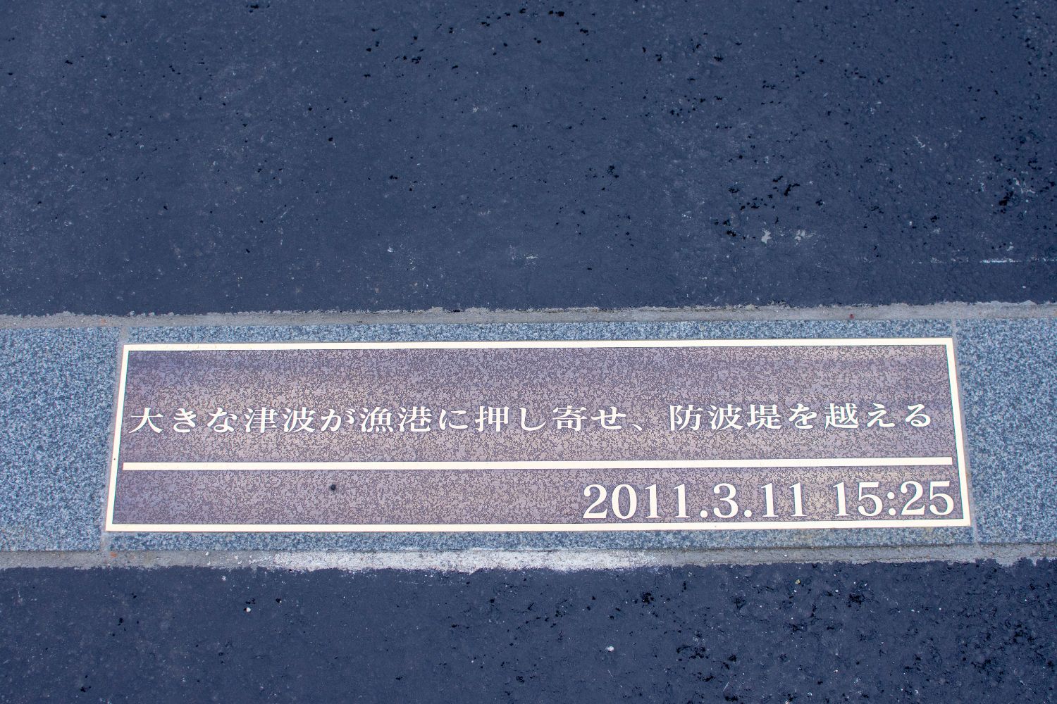 黒いアスファルトとグレーの縁石に囲まれ、「大きな津波が漁港に押し寄せ、防波堤を越える」「2011.3.11 15:25」と書かれた記念プレートの写真