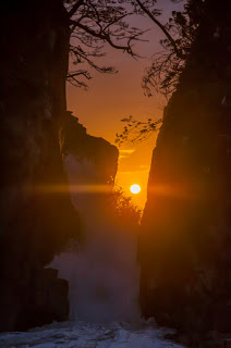 割れた岩の隙間から夕日がちょうど差し込み、岩の間から海に向かってまっすぐ沈んでいく神秘的な光景を写した夕暮れ時の神割崎の写真
