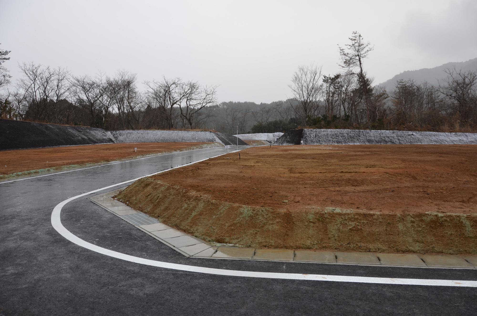 冬空の下、小雪が舞う中で舗装された黒いアスファルト道路が曲がりながら造成地の区画を縫うように伸び、周囲には土が整地された区画、そして葉を落とした木々が立ち並んでいる写真