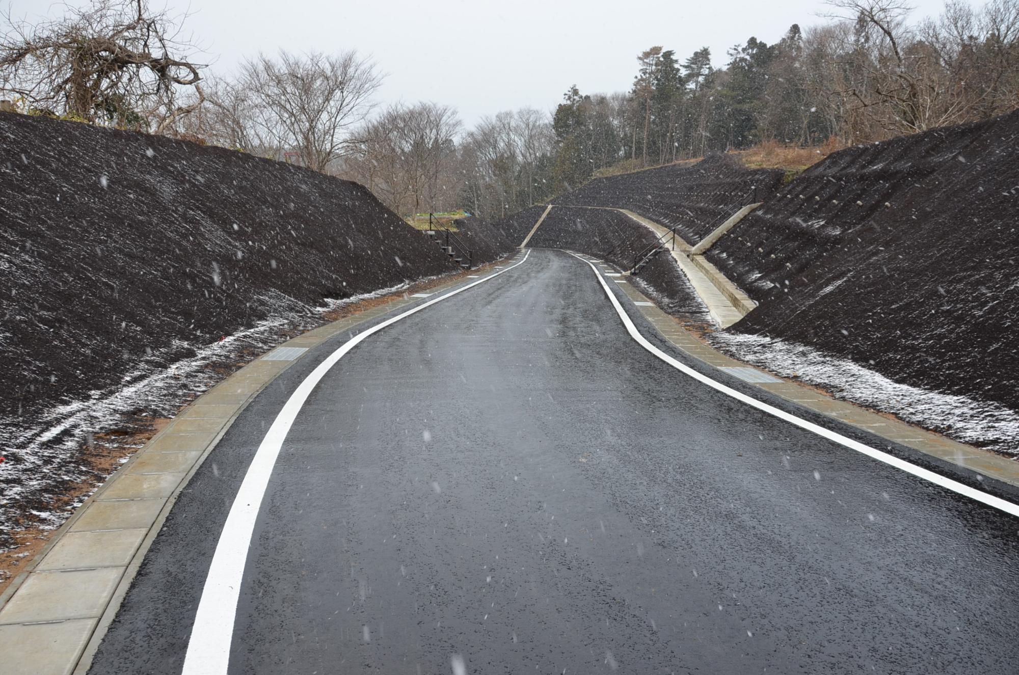 小雪が舞う中、黒く締め固められた法面に囲まれた新しい舗装道路が緩やかにカーブしながら奥へと続いており、右手には排水用の側溝と階段が設置されている写真
