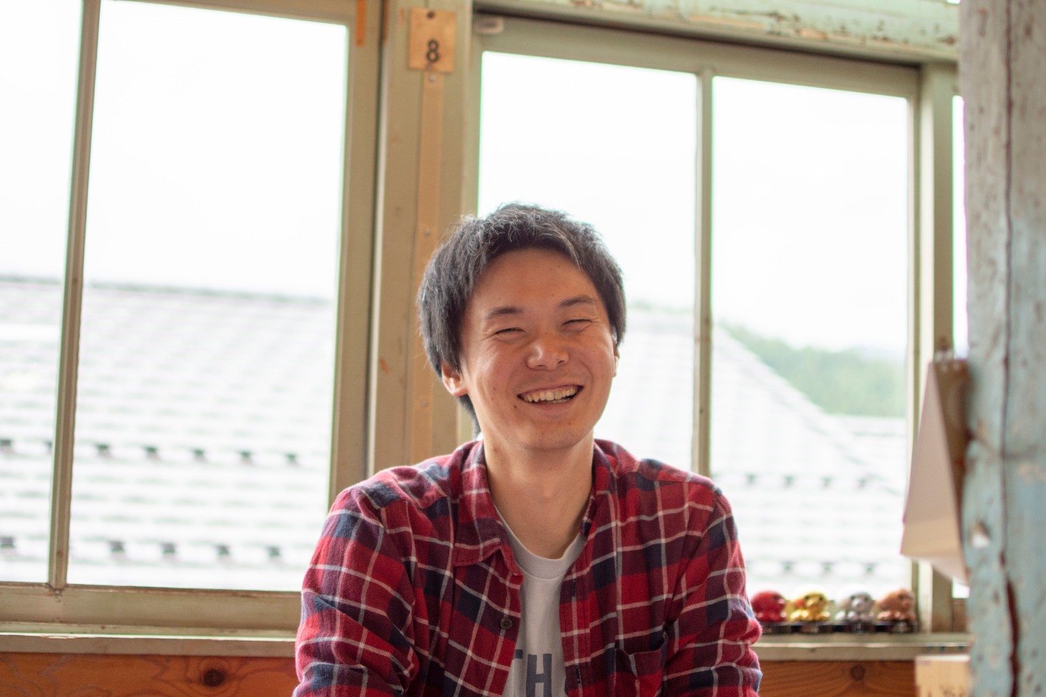 窓際で赤いチェック柄のシャツを着た大森さんが笑顔で座っている写真