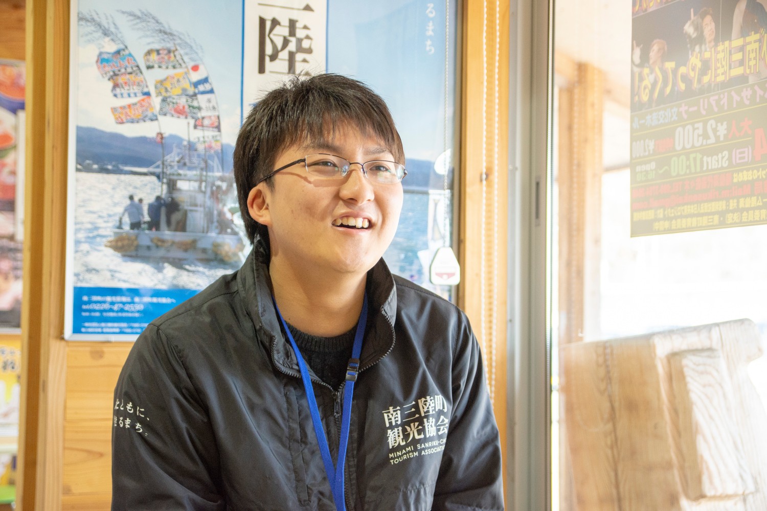 南三陸のポスターが壁に貼られた観光案内所内で、男性が椅子に座って笑顔で話している様子をアップで撮影した写真