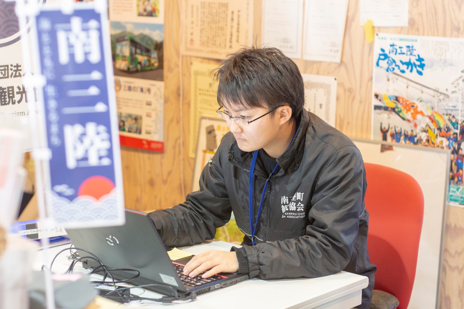 室内で「南三陸町観光協会」と書かれたジャンパーを着た男性が真剣な表情でパソコン作業をしている写真